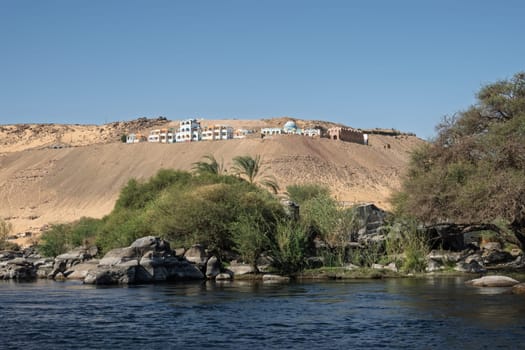 Granite rocks in the fast flowing water of the River Nile near Aswan Egypt with resort on hilltop