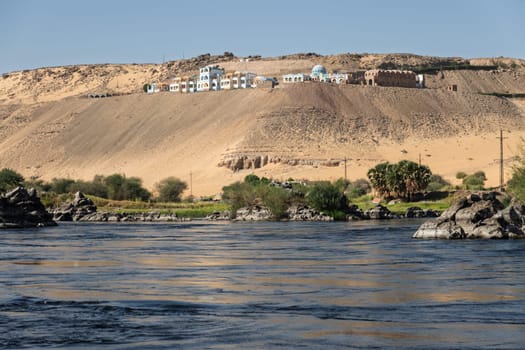 Granite rocks in the fast flowing water of the River Nile near Aswan Egypt with resort on hilltop