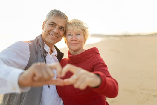 Senior couple making heart shape with hands, enjoying love and companionship on beach during sunset