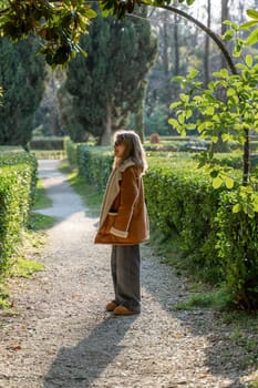 Woman in brown sheepskin coat stands on a sunlit path between neatly trimmed hedges in a park. Autumnal foliage surrounds.
