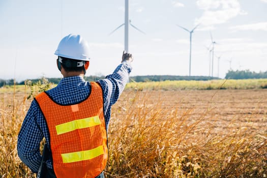 An Asian engineer helmet-wearing inspects windmill farm. Clean energy generation assured focusing on turbine efficiency. Global innovation a portrait of expertise in electric security.