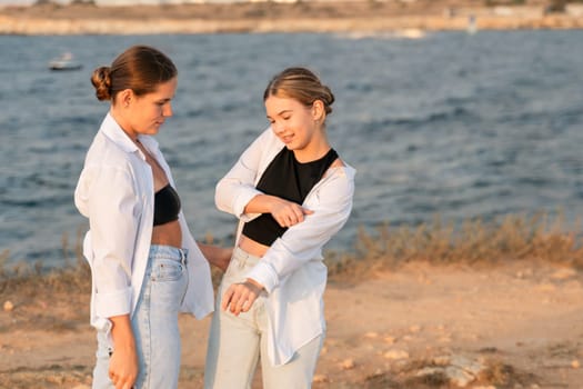 Two young women stand on a beach, one in a white shirt and jeans, the other in a black crop top and jeans. They are laughing and talking to each other.