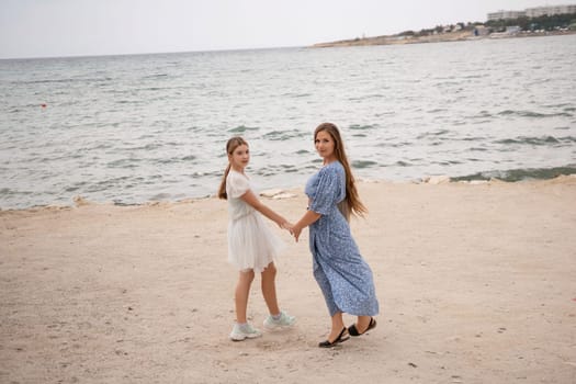 Two women, holding hands, stand on a sandy beach near a calm sea. One wears a white dress, the other a blue one. They are smiling.