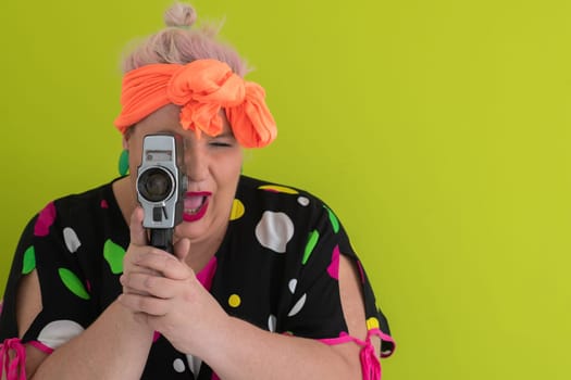 Playful and plus size woman with pink hair and orange headband aiming a vintage film camera, wearing a colorful polka dot dress against a vivid green background.