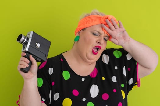 Playful and plus size woman with pink hair and orange headband aiming a vintage film camera, wearing a colorful polka dot dress against a vivid green background.