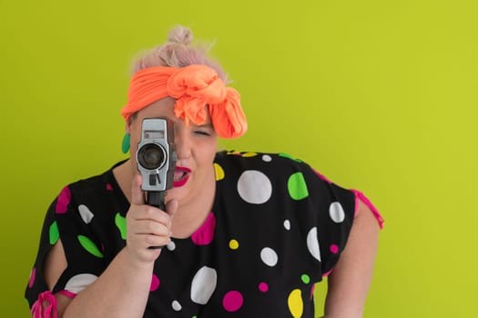 Playful and plus size woman with pink hair and orange headband aiming a vintage film camera, wearing a colorful polka dot dress against a vivid green background.