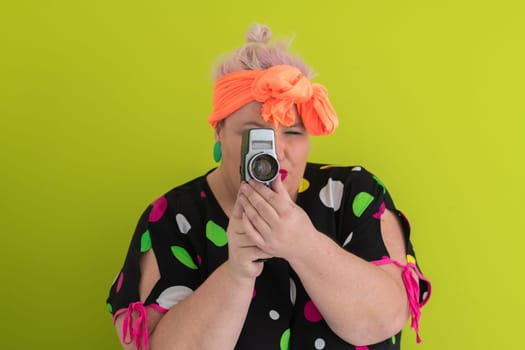 Playful and plus size woman with pink hair and orange headband aiming a vintage film camera, wearing a colorful polka dot dress against a vivid green background.