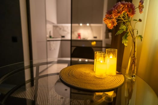 LED candlelight illuminates a glass-encased centerpiece on a round table, with blurred kitchen and flowers in the background. Warm, inviting ambiance.