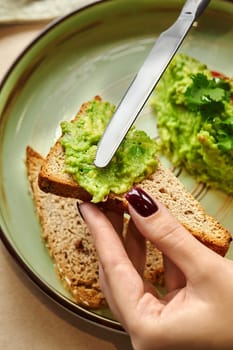 Woman's hand spreading mashed avocado on a slice of whole wheat toast, preparing a healthy and nutritious breakfast or snack
