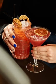 Two female hands holding red cocktails decorated with dehydrated orange slices and ice cubes