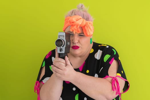 Playful and plus size woman with pink hair and orange headband aiming a vintage film camera, wearing a colorful polka dot dress against a vivid green background.