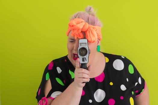 Playful and plus size woman with pink hair and orange headband aiming a vintage film camera, wearing a colorful polka dot dress against a vivid green background.