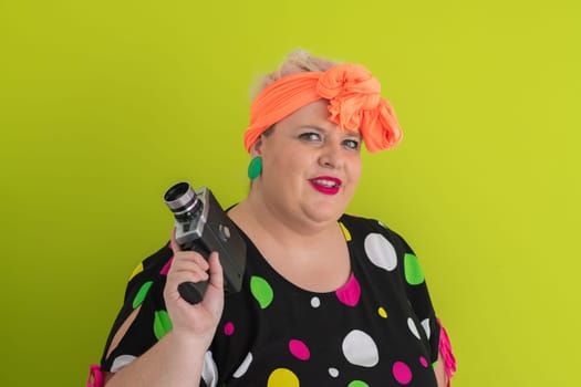 Playful and plus size woman with pink hair and orange headband aiming a vintage film camera, wearing a colorful polka dot dress against a vivid green background.