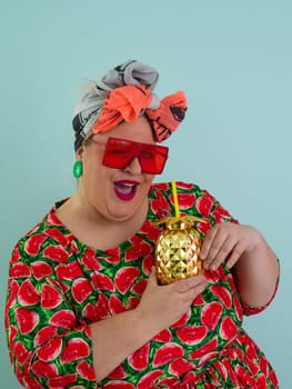 Eccentric plus size woman in a watermelon-patterned dress and colorful headwrap holding a golden pineapple-shaped cup with a straw, playfully glancing over her red sunglasses.