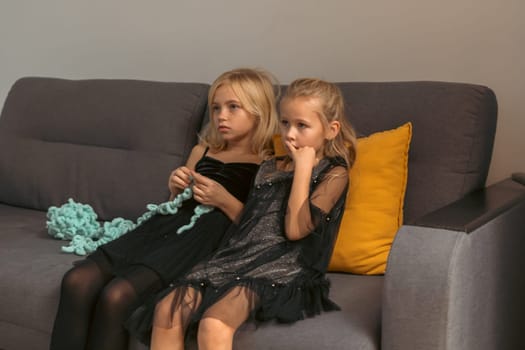 Two girls in black dresses sit on a gray couch. One knits with teal yarn; the other watches. A mustard-yellow pillow rests nearby.