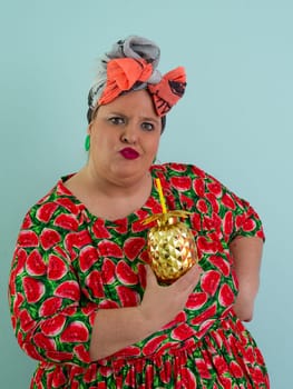 Eccentric plus size woman in a watermelon-patterned dress and colorful headwrap holding a golden pineapple-shaped cup with a straw, playfully glancing over her red sunglasses.
