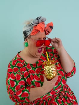 Eccentric plus size woman in a watermelon-patterned dress and colorful headwrap holding a golden pineapple-shaped cup with a straw, playfully glancing over her red sunglasses.