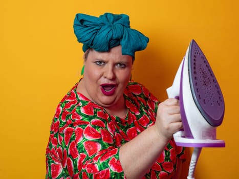 Plus size woman in a vibrant watermelon-patterned dress and teal headwrap, holding an electric iron and shouting with a humorous expression against a bright orange background. Conceptual and colorful portrait representing loudness, domestic chores, and quirky energy.