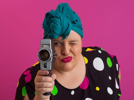 Playful and plus size woman with pink hair and orange headband aiming a vintage film camera, wearing a colorful polka dot dress against a pink background.
