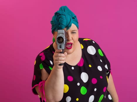 Playful and plus size woman with pink hair and orange headband aiming a vintage film camera, wearing a colorful polka dot dress against a pink background.