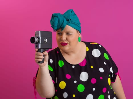 Playful and plus size woman with pink hair and orange headband aiming a vintage film camera, wearing a colorful polka dot dress against a pink background.