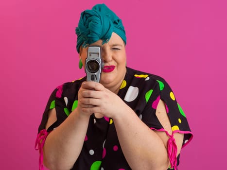 Playful and plus size woman with pink hair and orange headband aiming a vintage film camera, wearing a colorful polka dot dress against a pink background.