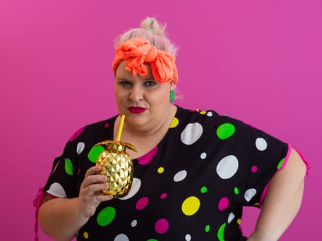 Confident plus size woman in vibrant retro outfit holding a golden pineapple drink cup while posing with attitude against a bright pink background.