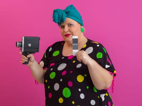 Playful and plus size woman with pink hair and orange headband aiming a vintage film camera, wearing a colorful polka dot dress against a pink background.