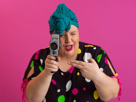 Playful and plus size woman with pink hair and orange headband aiming a vintage film camera, wearing a colorful polka dot dress against a pink background.