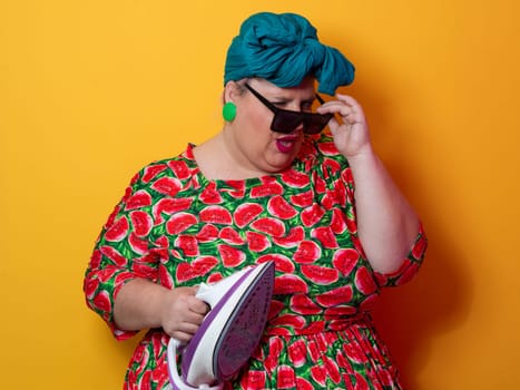 Plus size woman in a vibrant watermelon-patterned dress and teal headwrap, holding an electric iron and shouting with a humorous expression against a bright orange background. Conceptual and colorful portrait representing loudness, domestic chores, and quirky energy.