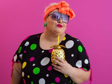 Confident plus size woman in vibrant retro outfit holding a golden pineapple drink cup while posing with attitude against a bright pink background.