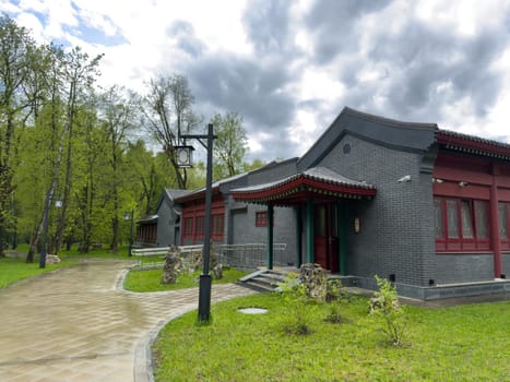Moscow, Russia, April 25, 2025: Pathway leading past traditional Chinese style building in Huamin Park with gray tiled walls, red wooden elements and green surroundings under dramatic sky