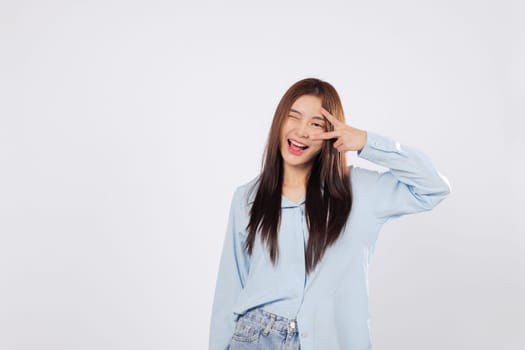 Victory gesture near eye with smile, showing peace and joy. Victory mood captured isolated on white background, optimistic Asian woman expresses positive vibes and excitement.