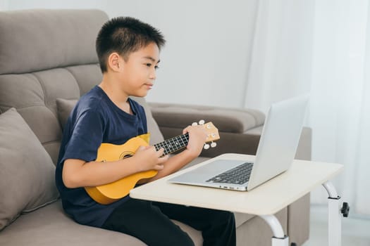 Boy learning ukulele on a sofa while watching a tutorial on a laptop. The cozy living room scene highlights the integration of technology and creative education in a modern home environment.