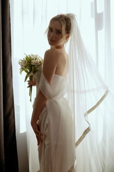 blonde bride in a long veil stands in a long white dress near the window with a bouquet of lilies in her hands. Minimalistic interior, hotel or house. bride in a long dress with a slit on the side