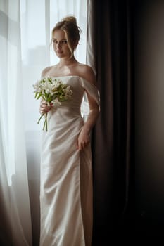 Beautiful blonde bride standing in a long white dress near the window with a bouquet in her hands. Minimalistic interior, hotel or house. Bride in a long dress with a slit on the side