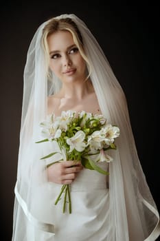 Portrait of bride with a lush veil, in a white dress near the window with a bouquet of lilies. Minimalistic interior, dark background, natural lighting. Bride with makeup and a high hairstyle,.