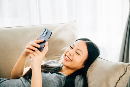 Young Asian woman relaxes on sofa texting on smartphone. Enjoying relaxation chatting and online shopping. Modern technology for communication and connection.