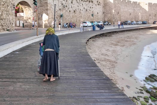 Crete.Greece - may 17, 2025: An elderly woman pushes a cart along a wooden path near a historic city wall and beach, capturing a moment of daily life in a picturesque setting.