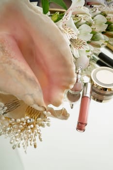 Lip gloss, highlighter, powder, flowers, hair ornaments with pearls, makeup brush, big seashell on mirror background. flat lay with space for text. vertical photo, Cosmetic composition