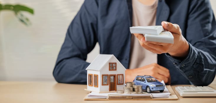 Money bills held above house and car models with paperwork. Represents tax-saving strategies, cash flow, and property loan planning for financial stability and asset acquisition.