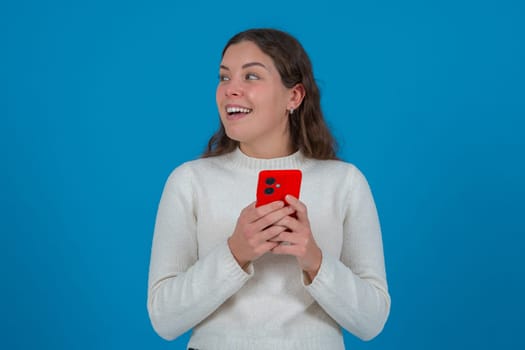 Happy young woman holding a red smartphone while looking to the side with a surprised expression. She stands in front of a bright blue background, expressing excitement and curiosity.