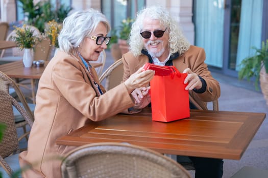 Elderly couple wearing glasses and brown coats enjoying coffee at a cozy outdoor cafe. They share a warm moment, smiling and relaxed in a peaceful, casual atmosphere during daytime. Vertical