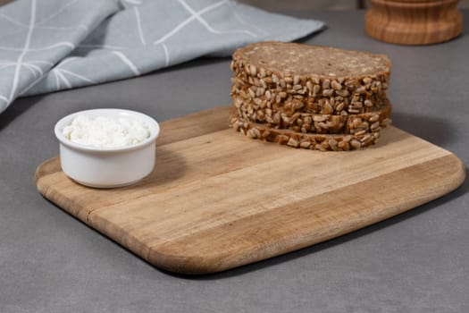 Three hearty slices of whole grain bread topped with sunflower seeds sit on a wooden cutting board alongside a bowl of creamy cottage cheese, creating a nutritious meal option.