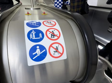 Escalator safety instruction sticker with visible passengers in background, showing real life context of public transport and importance of following safety guidelines