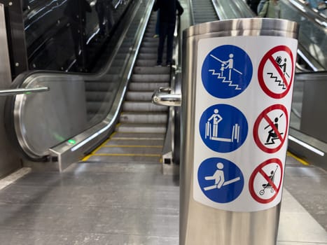 Closeup of escalator safety instruction sign in metro station with visible stairs and people in background, illustrating public space design and visual safety communication