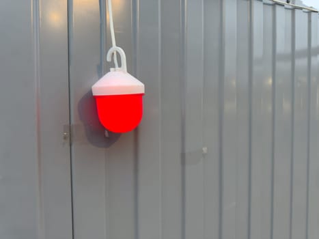 Single red warning light hanging on gray metal construction fence, glowing brightly in dim ambient light. Image highlights urban safety equipment and industrial aesthetics in public environments