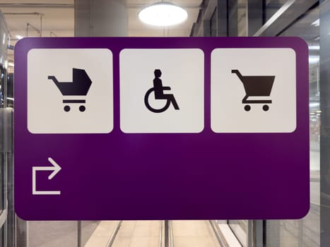 Minimalistic wayfinding sign with icons on purple background, showing designated zones for parents with strollers, wheelchair users and shopping carts near entrance of modern commercial building