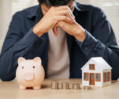 Worried man with a piggy bank, coin stacks, and house model nearby. Represents financial risks, property problems, money struggles, and the emotional toll of budgeting and unhappy banking situations.