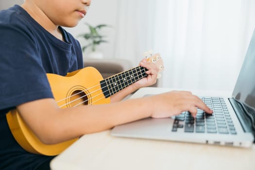Hands of a kid boy playing a ukulele, with a laptop nearby showing an online music course. The living room scene creates a cozy and educational environment for his practice and creativity.